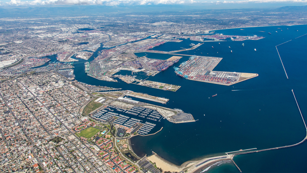 A high-altitude aerial view of the Los Angeles and Long Beach port complex, showing massive container terminals, breakwaters, and the surrounding city.