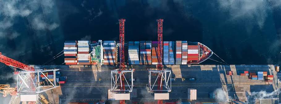 A top-down aerial view of a container ship docked at a port, loaded with colorful shipping containers and serviced by tall red gantry cranes.