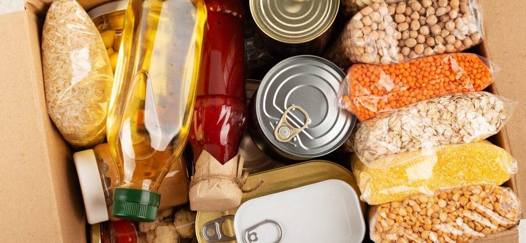 An overhead view of a cardboard box filled with various food items, including bags of rice, oats, lentils, a bottle of oil, and canned goods.